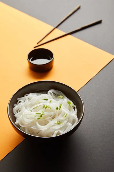 Rice noodles in bowl near chopsticks and soy sauce on yellow and black surface — Stock Photo