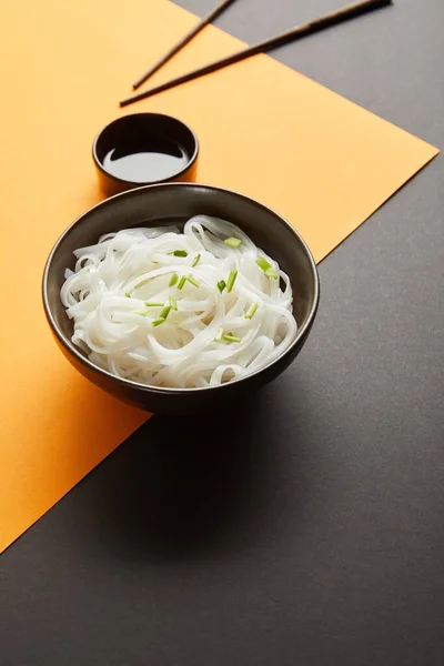 Rice noodles in bowl near chopsticks and soy sauce on yellow and black surface — Stock Photo