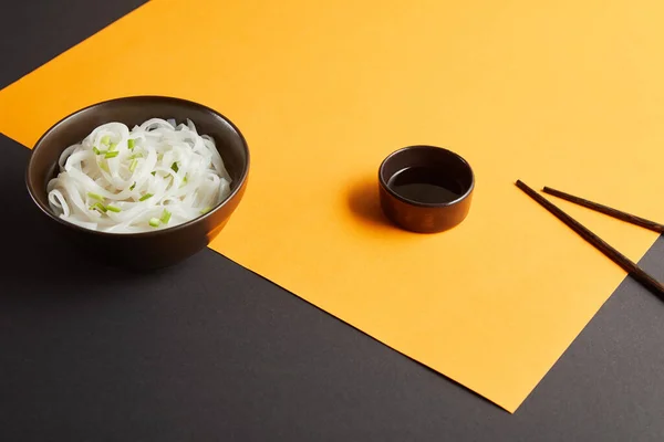 Rice noodles in bowl near chopsticks and soy sauce on yellow and black surface — Stock Photo