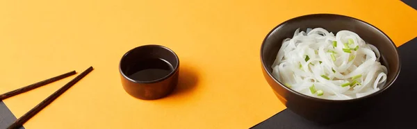 Rice noodles in bowl near chopsticks and soy sauce on yellow and black surface, panoramic shot — Stock Photo
