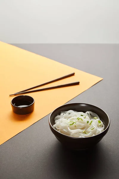 Selective focus of rice noodles in bowl near chopsticks and soy sauce on yellow and black surface isolated on grey — Stock Photo