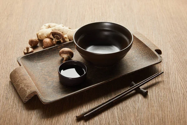 Empty bowl near chopsticks, soy sauce, ginger root and mushrooms on wooden tray — Stock Photo