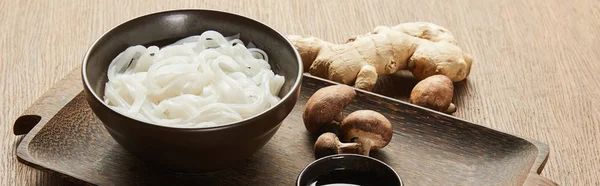 Rice noodles in bowl near soy sauce, ginger root and mushrooms on wooden tray, panoramic shot — Stock Photo