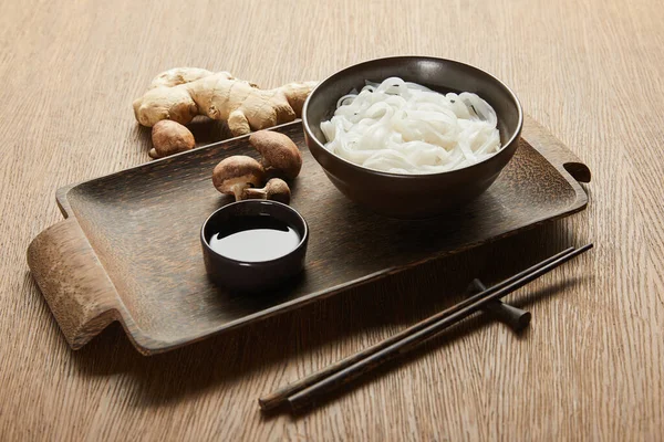 Tagliatelle di riso in ciotola vicino bacchette, salsa di soia, radice di zenzero e funghi su vassoio di legno — Foto stock