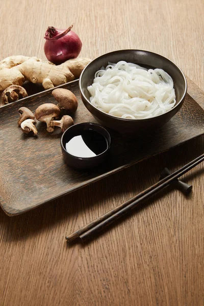 Рисовая лапша в миске возле палочек, соевый соус, корень имбиря, лук и грибы на деревянном подносе — стоковое фото