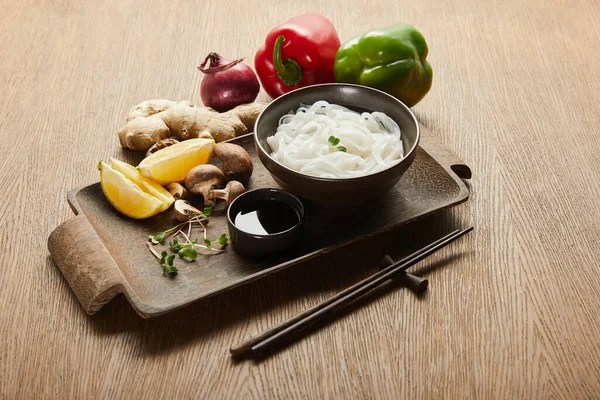 Nouilles de riz dans un bol près des baguettes, sauce soja, racine de gingembre, citron et légumes sur plateau en bois — Photo de stock