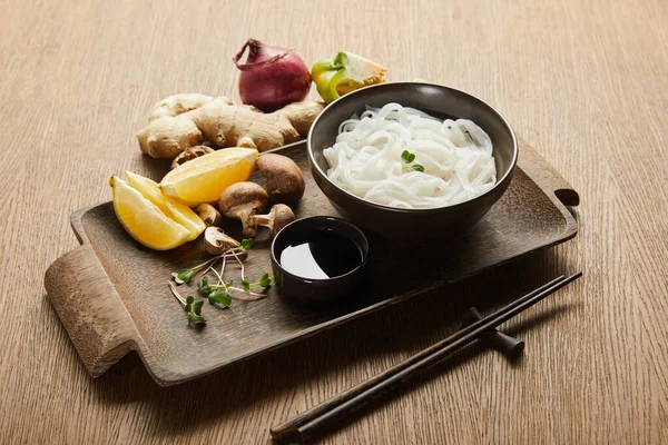 Rice noodles in bowl near chopsticks, soy sauce, ginger root, lemon and vegetables on wooden tray — Stock Photo