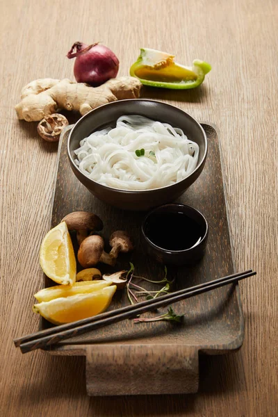 Рисовая лапша в миске возле палочек, соевый соус, корень имбиря, лимон и овощи на деревянном подносе — стоковое фото