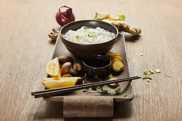 Fideos de arroz en cuenco cerca de palillos, salsa de soja, raíz de jengibre, limón y verduras en bandeja de madera. - foto de stock