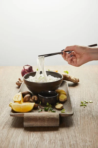 Cropped view of woman eating rice noodles with ginger root, lemon and vegetables on wooden tray isolated on grey — Stock Photo