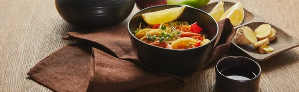 Nouilles aux crevettes, citron et légumes dans un bol près de sauce soja sur serviette sur table en bois, plan panoramique — Photo de stock