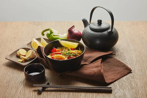 Noodles with shrimps and vegetables in bowl near chopsticks, soy sauce, lemon and ginger root, teapot on napkin on wooden table isolated on grey — Stock Photo