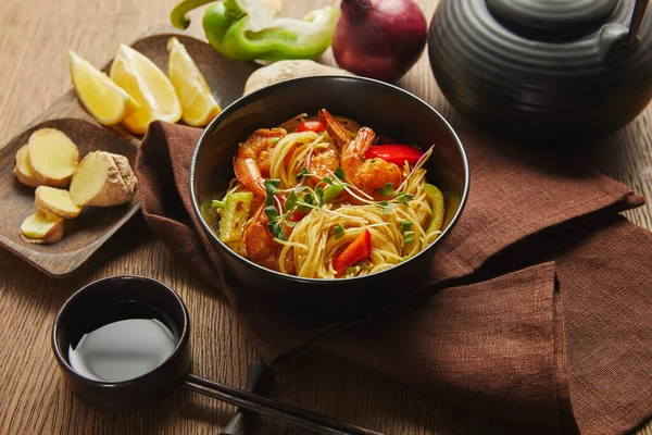 Noodles with shrimps and vegetables in bowl near chopsticks, soy sauce, lemon and ginger root, teapot on napkin on wooden table — Stock Photo