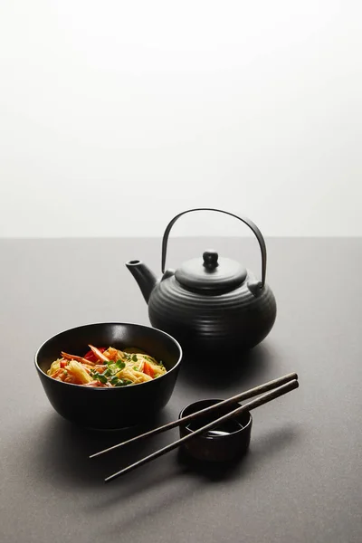Fideos con camarones y verduras en el cuenco cerca de limón, teapote, palillos de madera y salsa de soja en fondo negro aislados en gris. - foto de stock