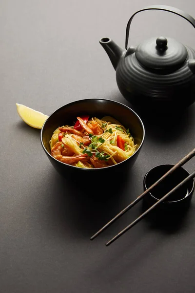 Noodles with shrimps and vegetables in bowl near lemon, teapot, wooden chopsticks and soy sauce on black background — Stock Photo