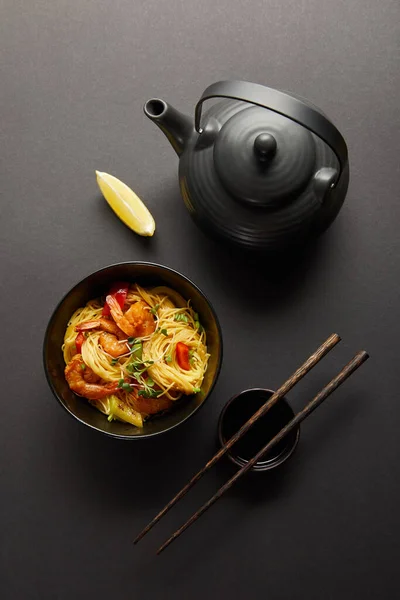Top view of noodles with shrimps and vegetables in bowl near lemon, teapot, wooden chopsticks and soy sauce on black background — Stock Photo