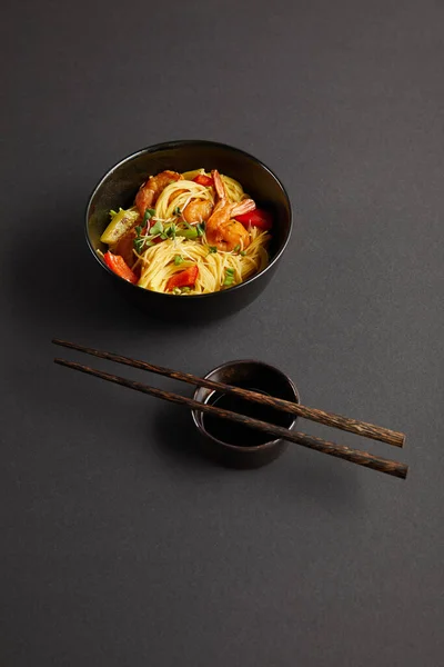 Noodles with shrimps and vegetables in bowl near wooden chopsticks and soy sauce on black background — Stock Photo