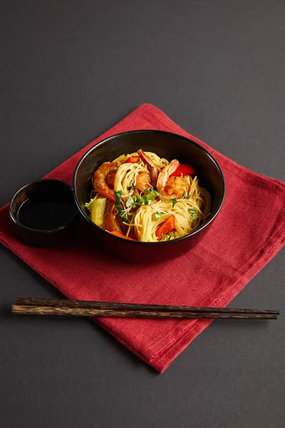 Noodles with shrimps and vegetables in bowl near wooden chopsticks, soy sauce on red napkin on black background — Stock Photo