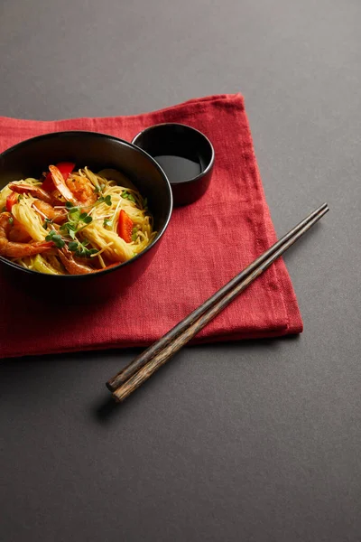Noodles with shrimps and vegetables in bowl near wooden chopsticks, soy sauce on red napkin on black background — Stock Photo