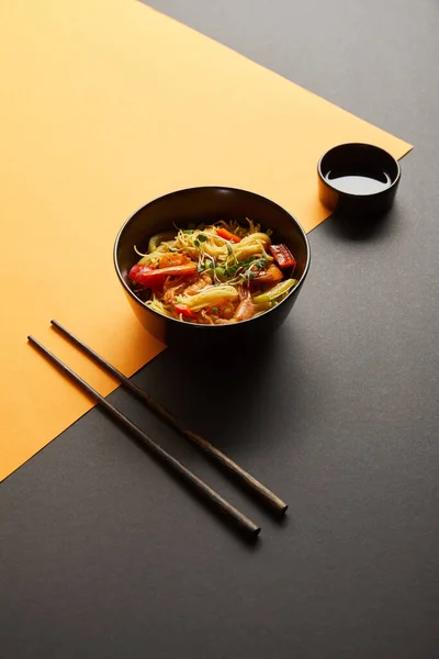 Tagliatelle con gamberetti e verdure in ciotola vicino bacchette, salsa di soia su sfondo nero e giallo — Foto stock