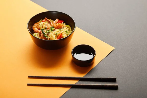 Nouilles aux crevettes et légumes dans un bol près des baguettes, sauce soja sur fond noir et jaune — Photo de stock