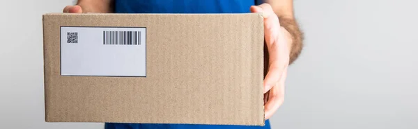 Cropped view of delivery man holding cardboard box with qr code and barcode isolated on grey, panoramic shot — Stock Photo