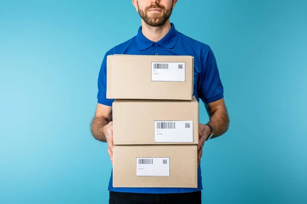 Vista recortada del mensajero sosteniendo paquetes de cartón con códigos qr y códigos de barras en tarjetas aisladas en azul - foto de stock