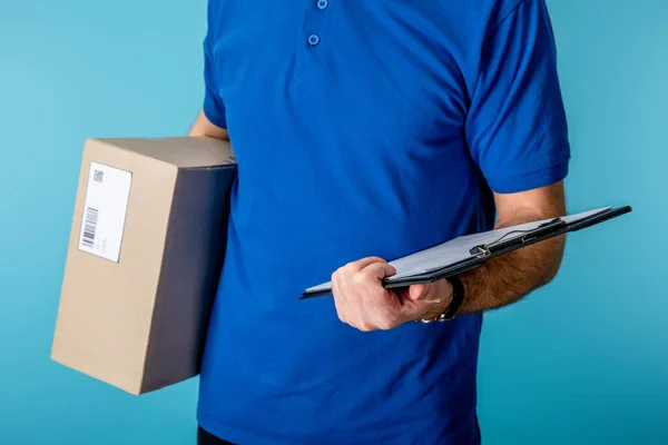 Cropped view of delivery man holding clipboard and cardboard package isolated on blue — Stock Photo