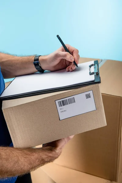 Cropped view of courier writing on clipboard with cardboard packages isolated on blue — Stock Photo