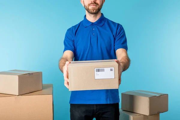 Vista recortada del mensajero que sostiene el paquete de cartón cerca de cajas aisladas en azul - foto de stock