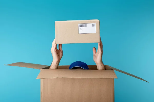 Vista cortada de correio em cap segurando pacote de papelão de caixa isolada em azul — Fotografia de Stock