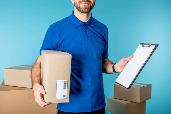 Vue recadrée du livreur tenant la boîte en carton et le presse-papiers avec espace de copie près des boîtes isolées sur bleu — Photo de stock