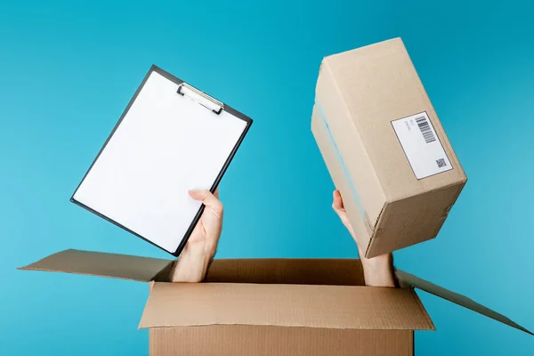 Manos de mensajero sujetando el portapapeles y el paquete de la caja de cartón aislado en azul - foto de stock