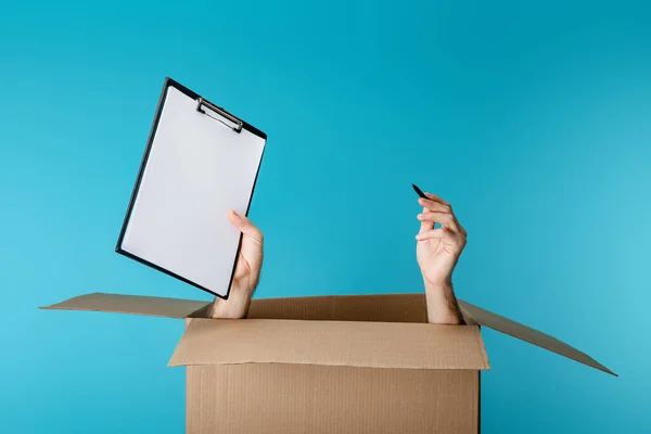 Manos del mensajero sujetando el portapapeles y la pluma de la caja de cartón aislada en azul - foto de stock