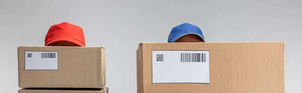 Panoramic shot of couriers in caps and cardboard packages with barcodes and qr codes on cards isolated on grey — Stock Photo