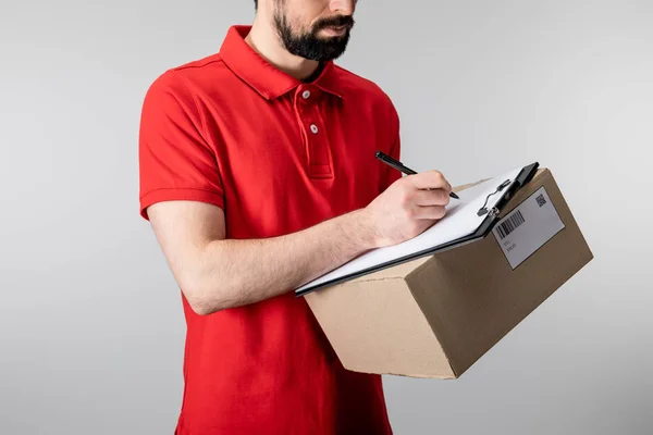 Vista recortada del repartidor escribiendo en portapapeles y sosteniendo la caja de cartón aislada en gris - foto de stock