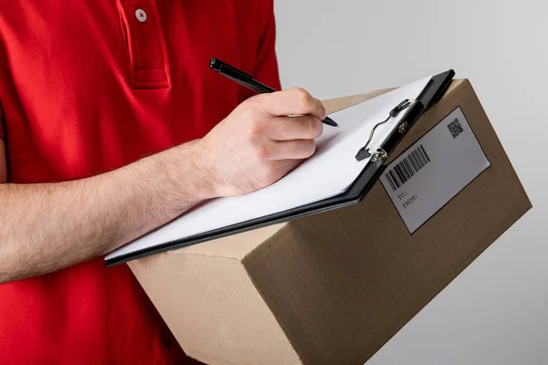 Cropped view of courier writing on clipboard with empty blank and holding cardboard box isolated on grey — Stock Photo