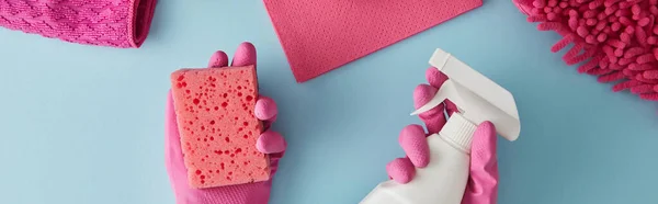 Panoramic shot of housekeeper in pink rubber gloves holding sponge and spray bottle on blue with rags — Stock Photo