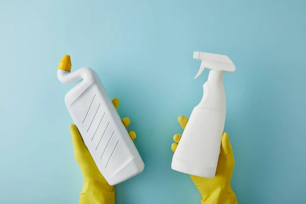 Cropped view of housekeeper in rubber gloves holding spray bottle and bottle with toilet cleaner on blue — Stock Photo