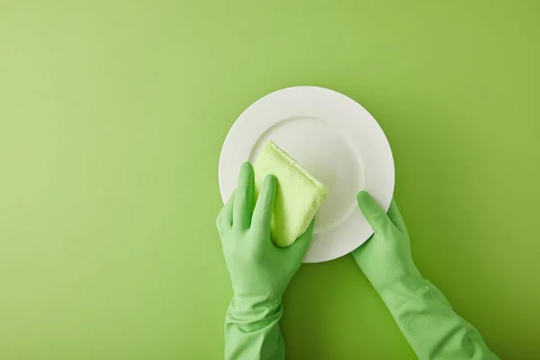 Обрезанный вид на домработницу в резиновых перчатках мыть тарелку с губкой на зеленый — стоковое фото