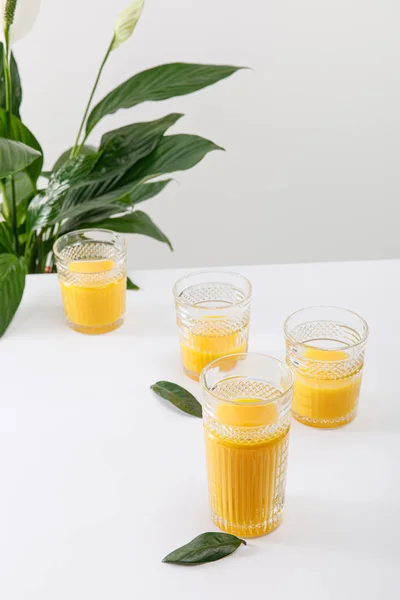 Copos de smoothie amarelo delicioso fresco na superfície branca perto de planta de lírio paz verde isolado em cinza — Fotografia de Stock