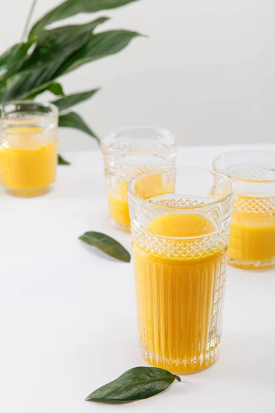 Selektiver Fokus von Gläsern mit frischem köstlichen gelben Smoothie auf weißer Oberfläche in der Nähe der grünen Friedenslilie-Pflanze isoliert auf grau — Stockfoto