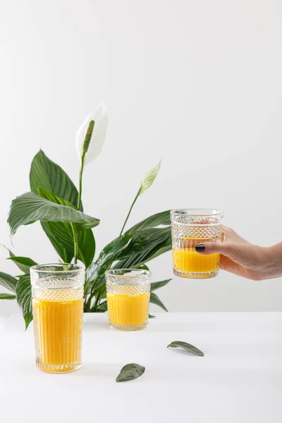 Vista cortada de mulher segurando vidro de smoothie amarelo delicioso fresco perto de planta de lírio paz verde isolado em branco — Fotografia de Stock