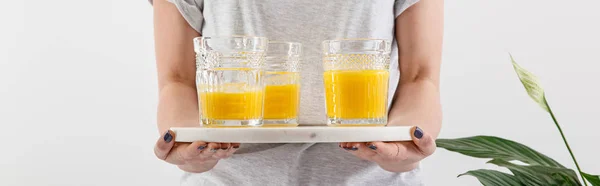 Cropped view of woman holding tray with glasses of fresh delicious yellow smoothie near green peace lily plant isolated on white, panoramic shot — Stock Photo