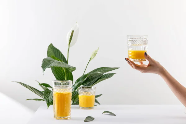 Vista recortada de la mujer sosteniendo el vaso de licuado amarillo delicioso fresco cerca de la planta de lirio de paz verde en blanco - foto de stock