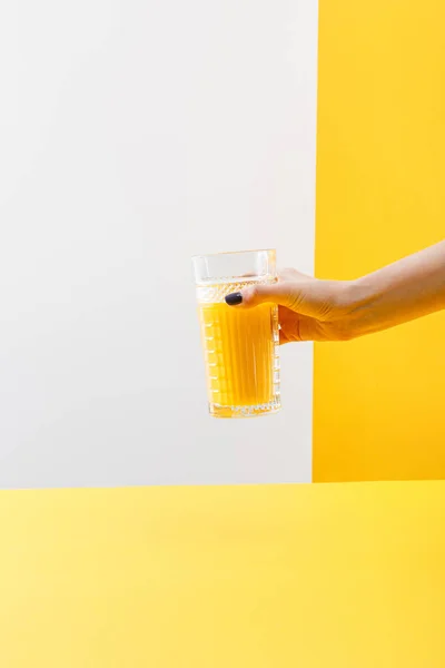 Vue recadrée d'une femme tenant un verre de délicieux smoothie jaune frais sur fond gris et jaune — Photo de stock