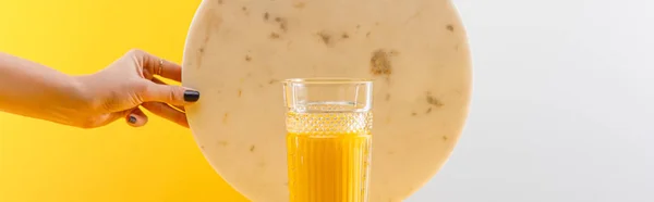 Vista cortada da mulher segurando círculo de mármore perto de vidro de smoothie amarelo delicioso fresco no fundo cinza e amarelo, tiro panorâmico — Fotografia de Stock