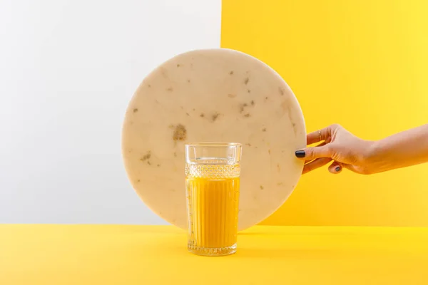 Vista recortada de la mujer sosteniendo círculo de mármol cerca del vaso de batido amarillo delicioso fresco sobre fondo gris y amarillo - foto de stock