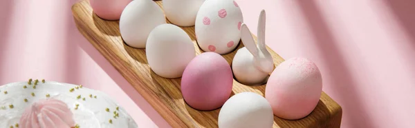 Oeufs de Pâques et lapin décoratif sur planche en bois près du gâteau sur fond rose, plan panoramique — Photo de stock