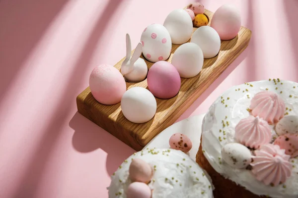 Concentration sélective des œufs de poulet et de caille peints, lapin décoratif sur planche de bois près de gâteaux de Pâques sur fond rose — Photo de stock